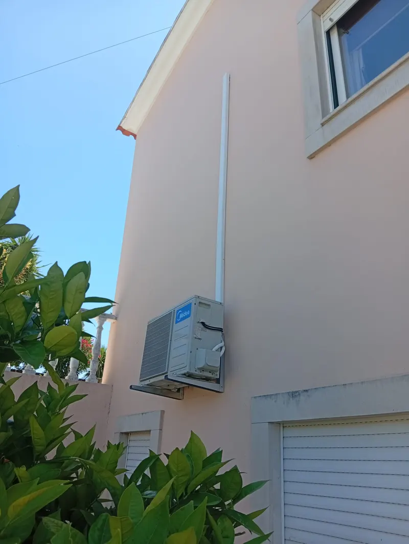 Unidade exterior de ar condicionado numa fachada branca em Cascais, com calha de tubagem até ao piso de cima.